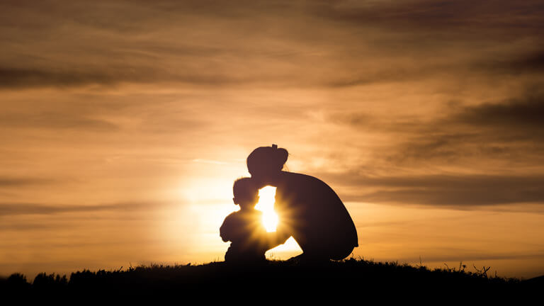 image of mother kneeling down in front of a sunset and kissing her small son on the head