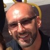 Close-up of smiling, balding man with glasses and facial hair, blurred background.