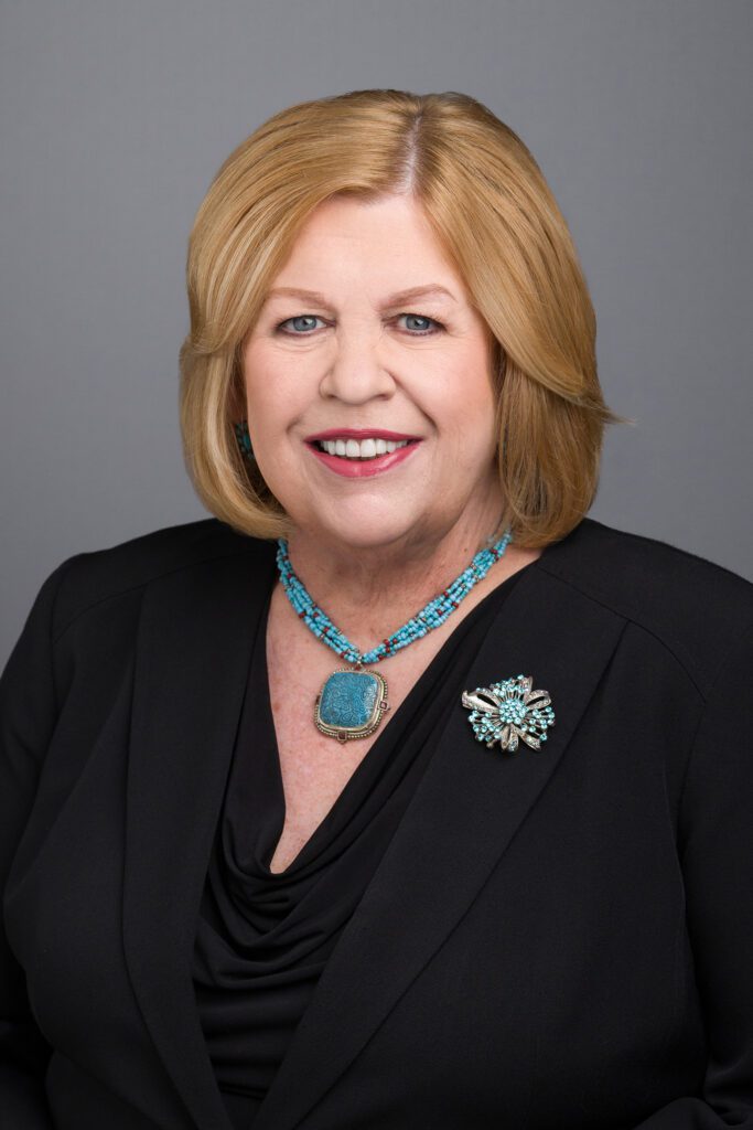 Professional portrait of Gayle Nathan, Esq., a distinguished attorney with short blonde hair, wearing a black suit, a turquoise necklace, and a decorative brooch. Gayle is smiling confidently, reflecting her experience and professionalism in the legal field.